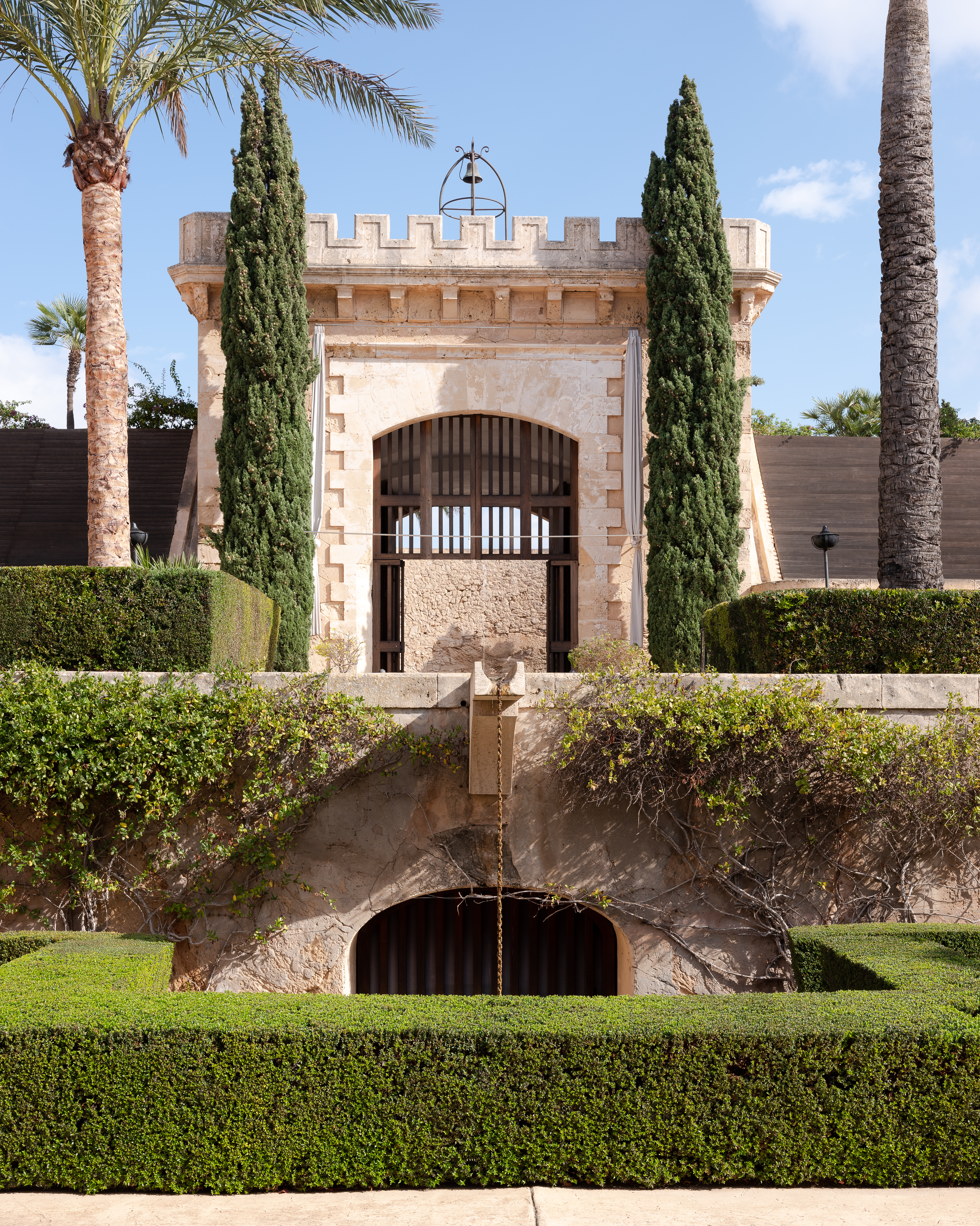 Stone building with gated archway surrounded by manicured hedges and palm trees under a sunny sky, evoking a Mediterranean garden.