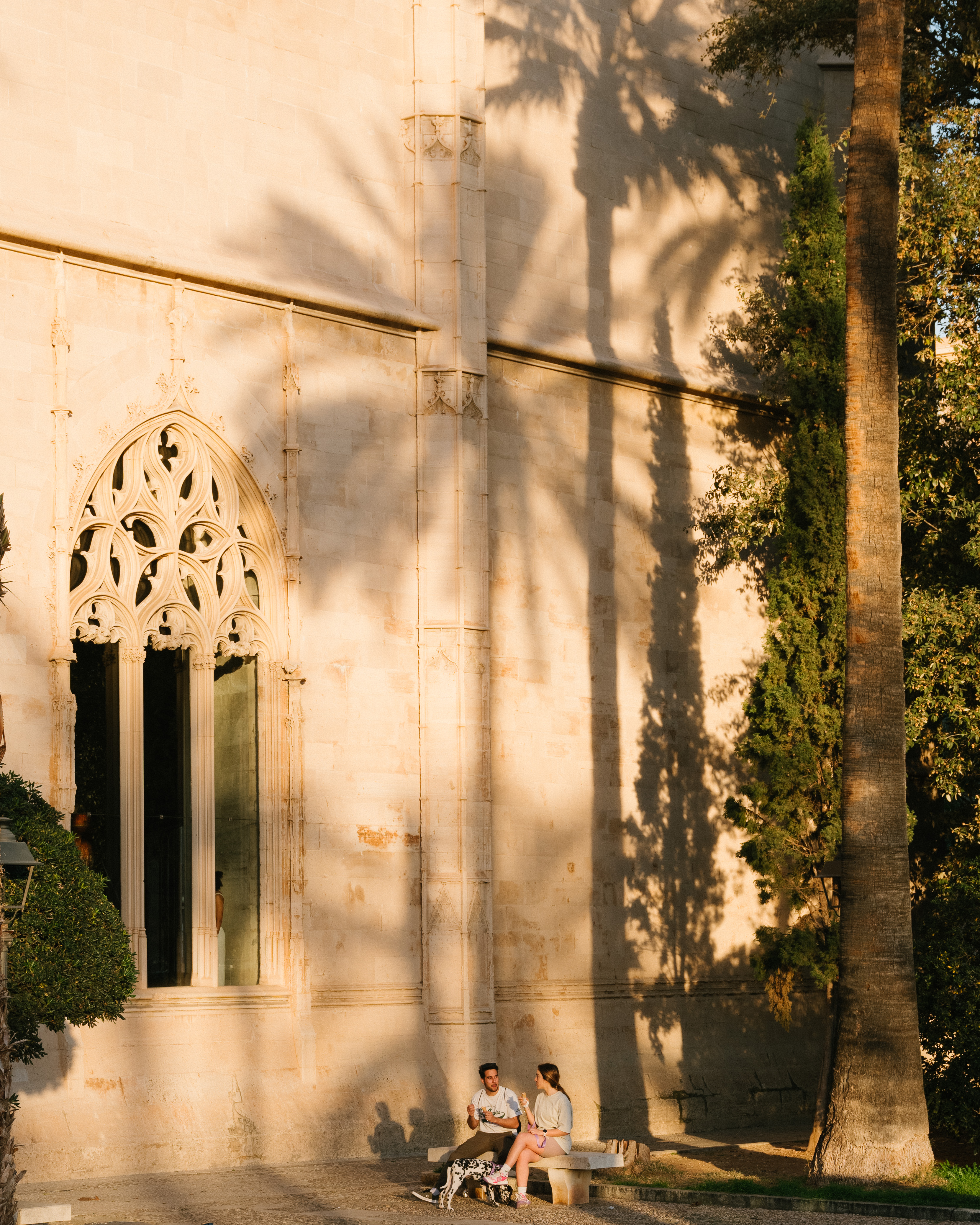 Couple sits on a bench near an ornate building while palm tree shadows fall on the stone wall in the afternoon sun.