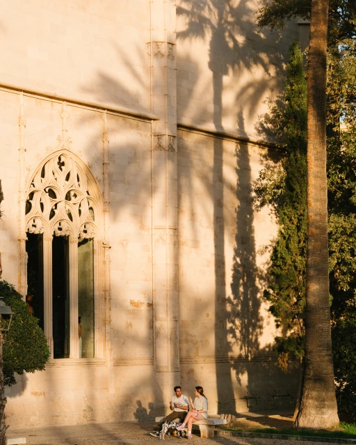 Couple sits on a bench near an ornate building while palm tree shadows fall on the stone wall in the afternoon sun.