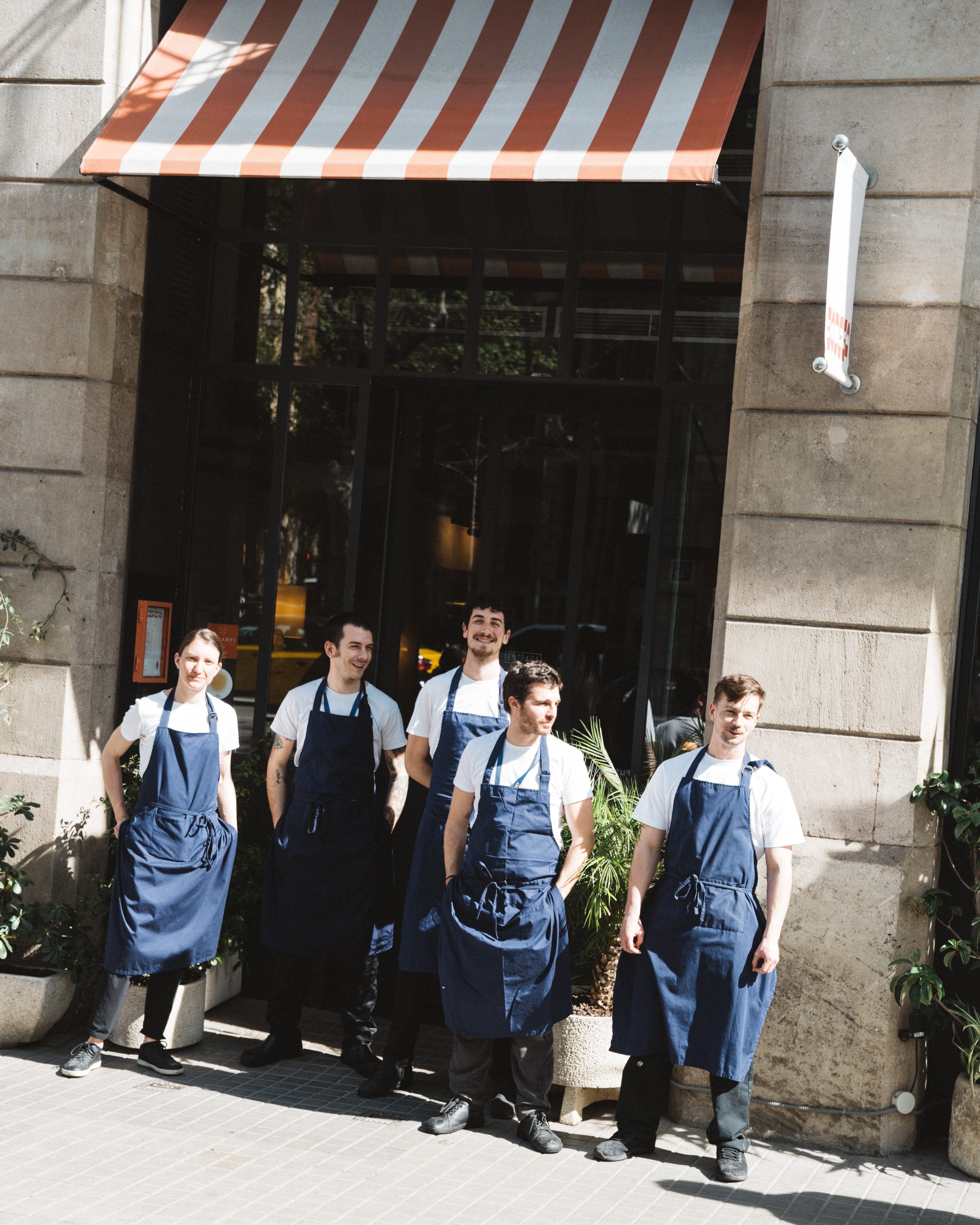 Group of five restaurant employees standing in uniform outside the establishment, with a striped awning in the background.