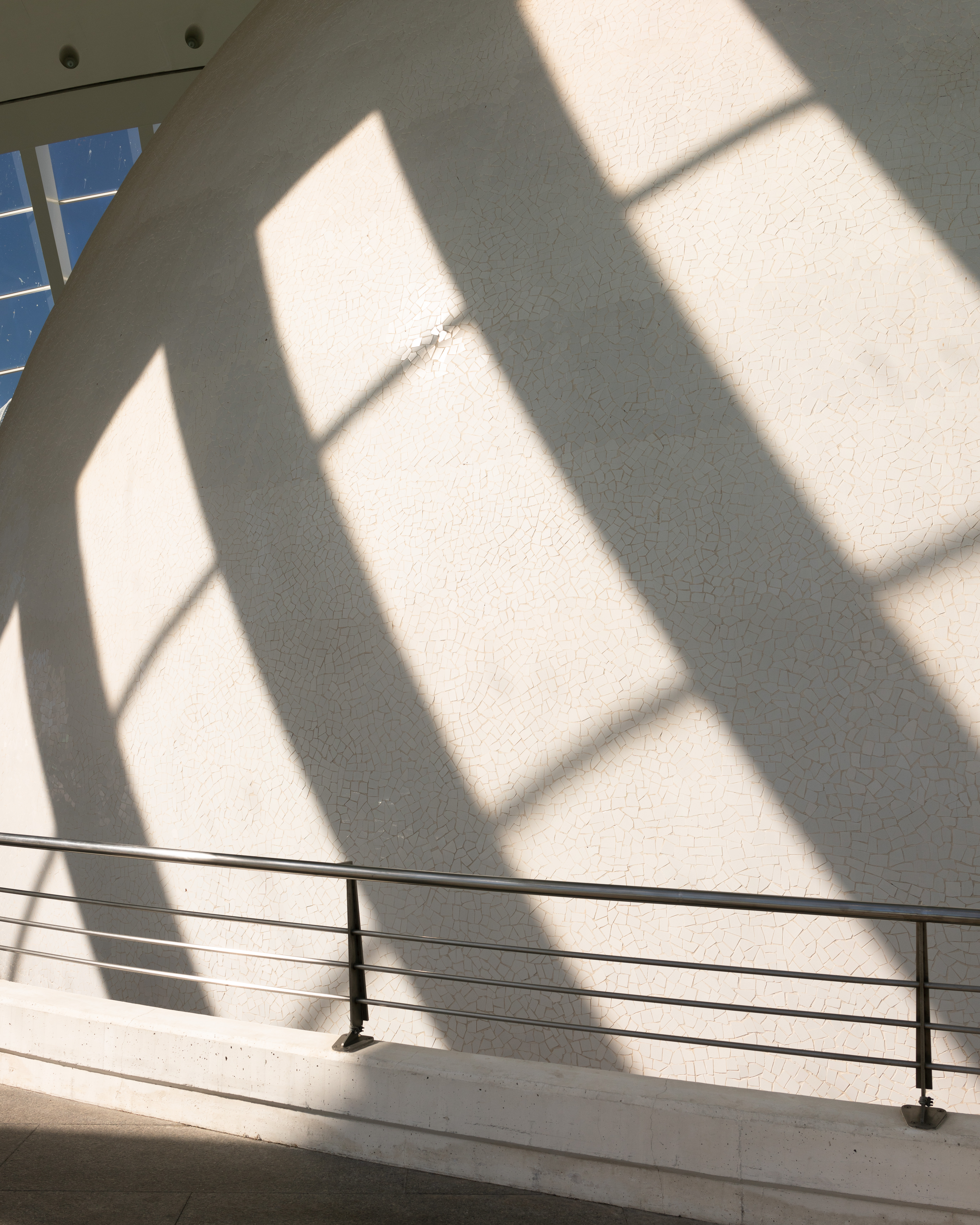 Hard geometric window shadows cast across a curved white textured wall above a metal railing in bright daylight.