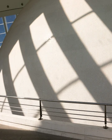 Hard geometric window shadows cast across a curved white textured wall above a metal railing in bright daylight.