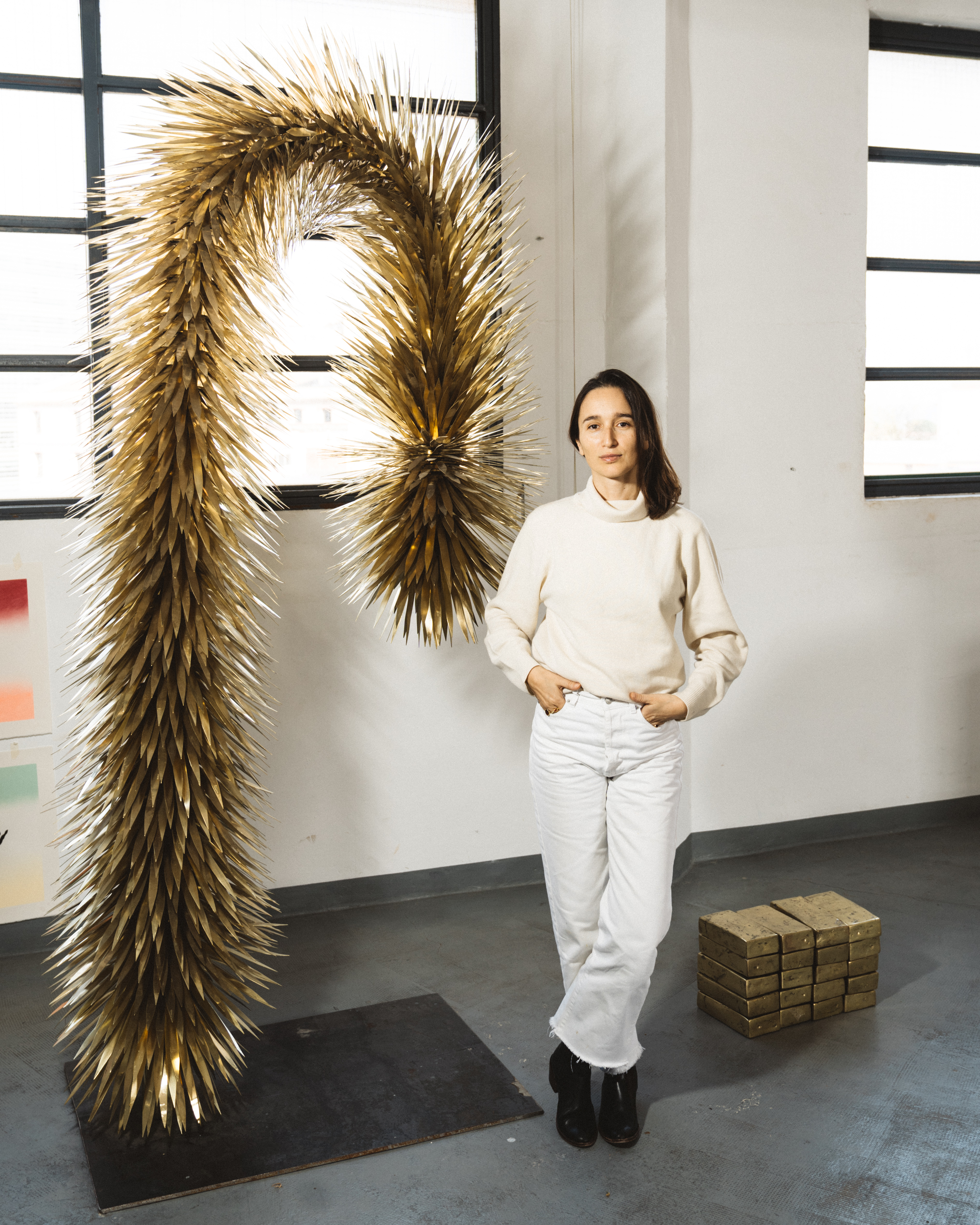 Woman in white next to a large, golden, letter ‘P’ sculpture made from spiky organic material in an industrial-style space.