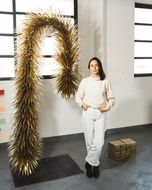 Woman in white next to a large, golden, letter ‘P’ sculpture made from spiky organic material in an industrial-style space.