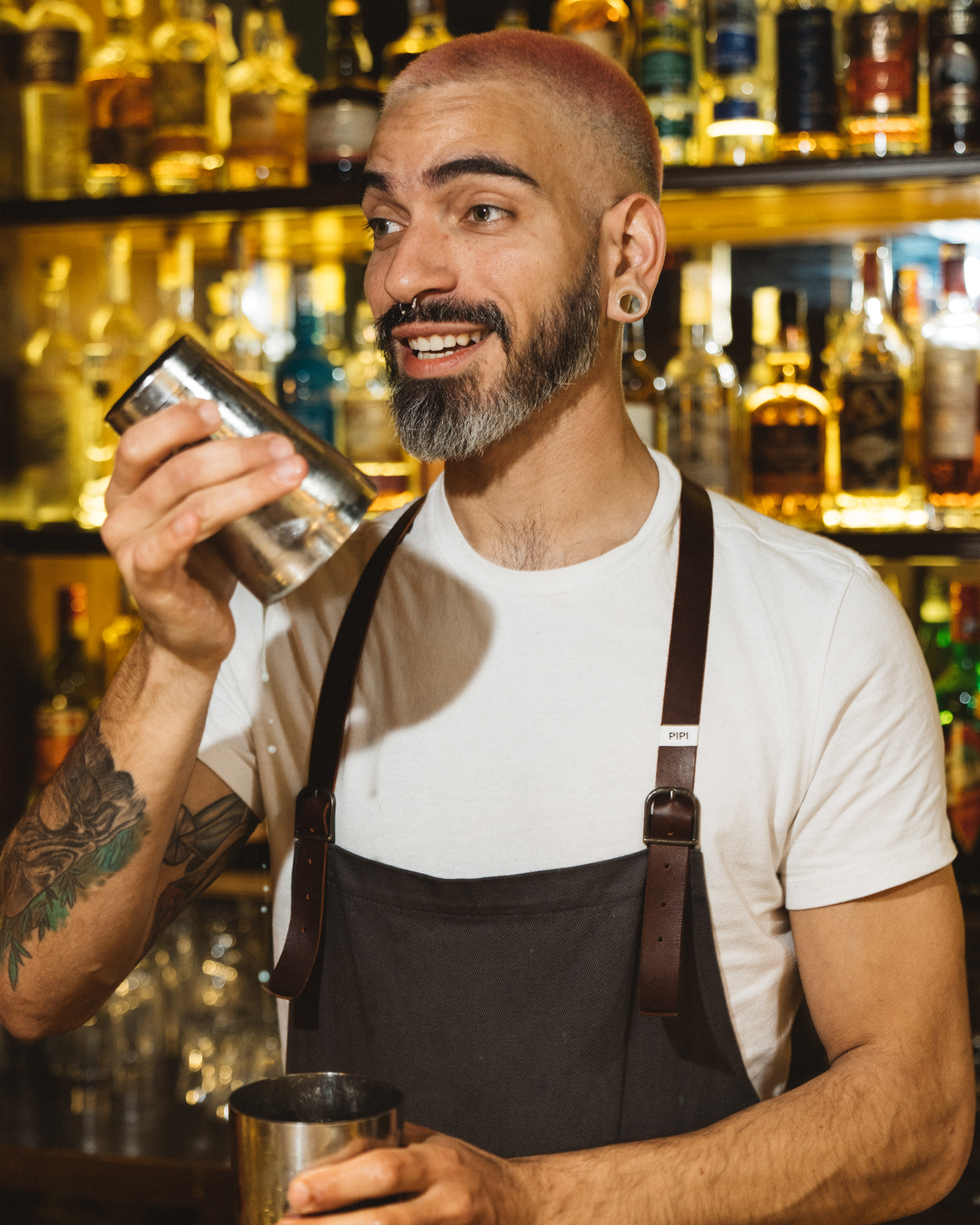 Bartender mixing a cocktail behind a bar. Adult male with beard and pink hair wearing an apron mixing a drink in a metal shaker.