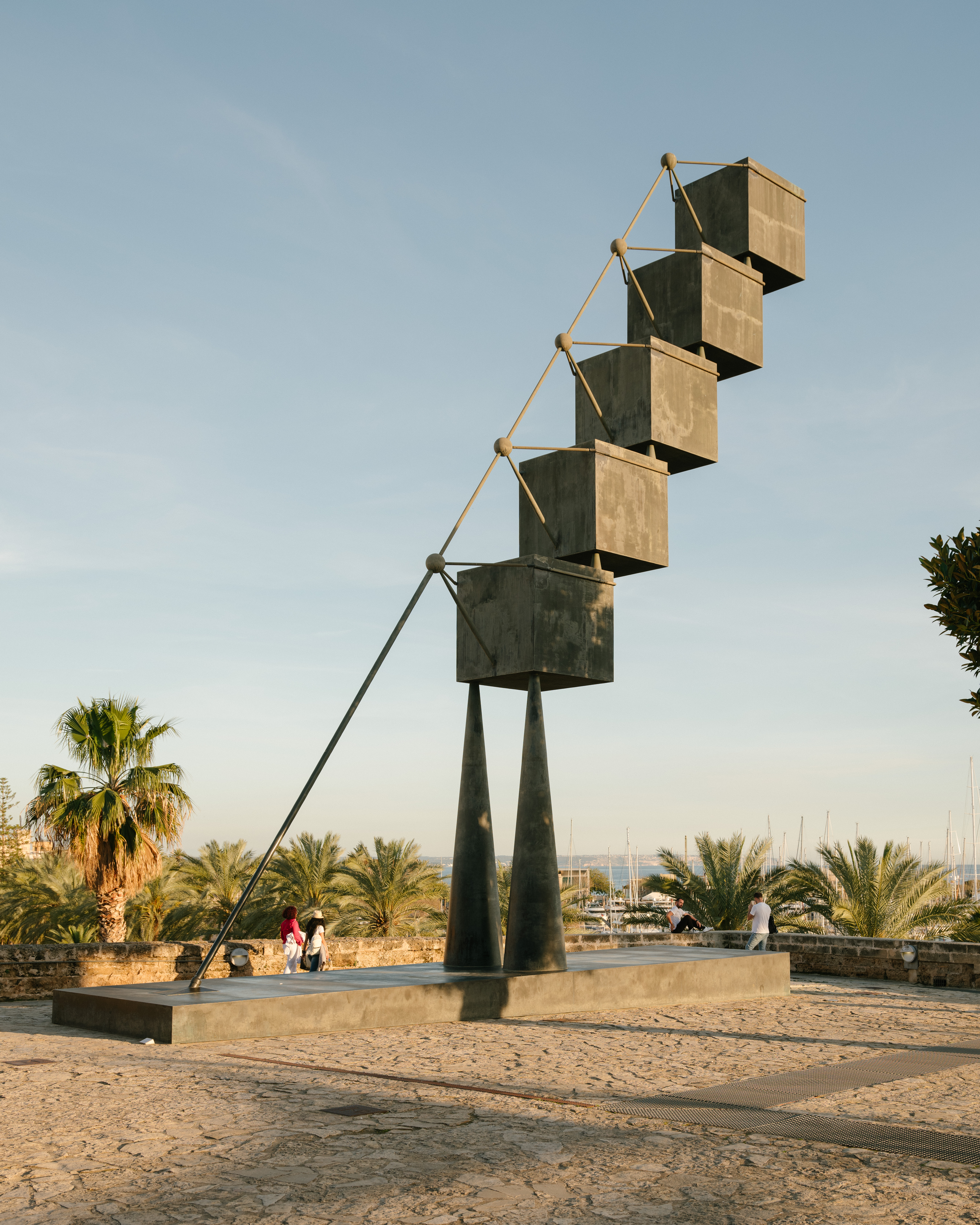Outdoor photo of a metal sculpture made of stacked cubes, set against a clear sky with trees and distant buildings in the background.