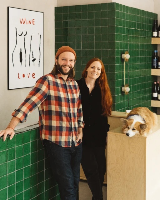 Couple and corgi dog in a wine shop with green tile wall and minimalist wine art. Stylish and inviting atmosphere for wine enthusiasts.