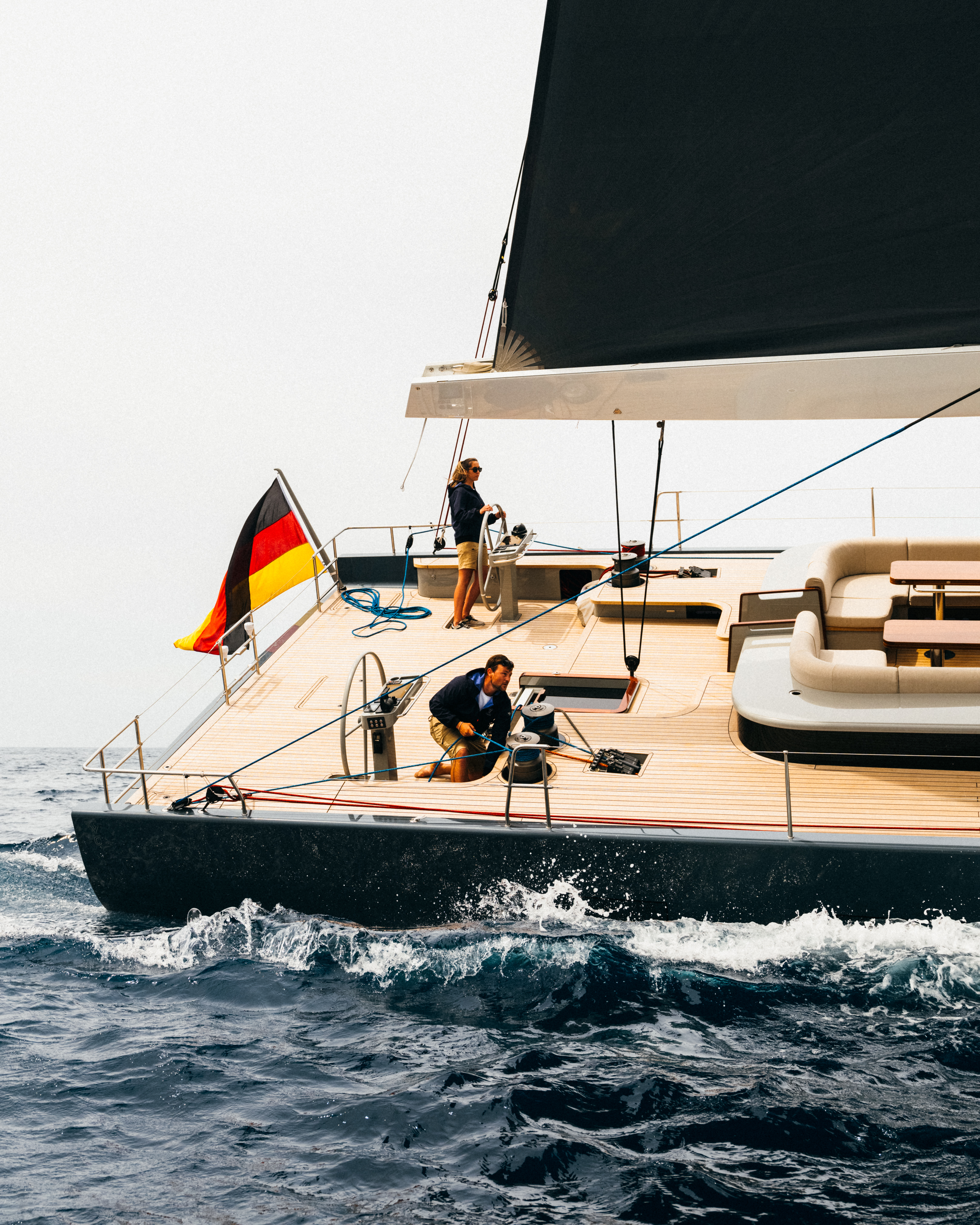 Two people sailing a modern yacht on the ocean, featuring a dark sail, wooden deck, and a German flag, under an overcast sky.