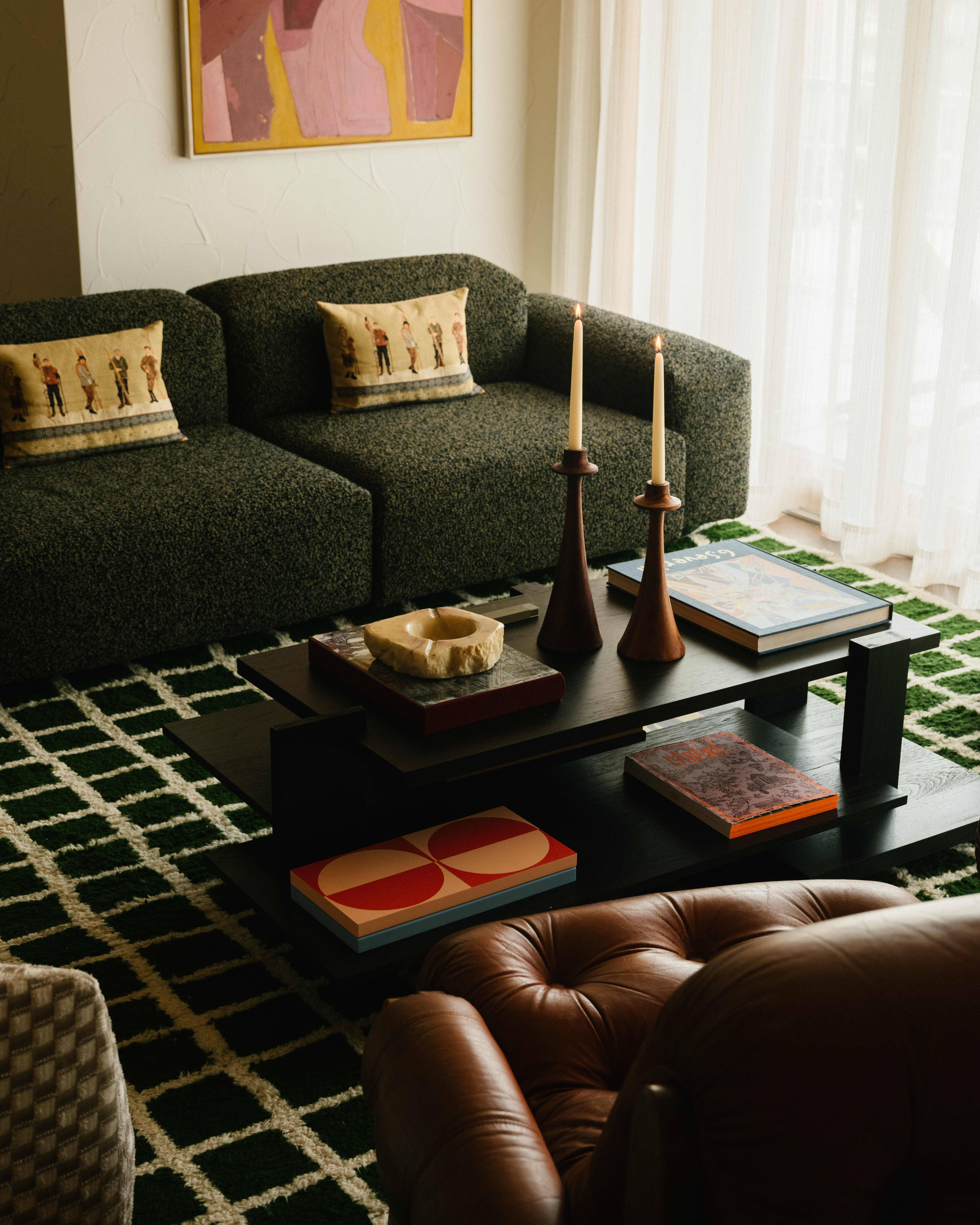 Interior view featuring a green couch with throw pillows, dark wood coffee table with decorative items, candles, books, and a green geometric rug.