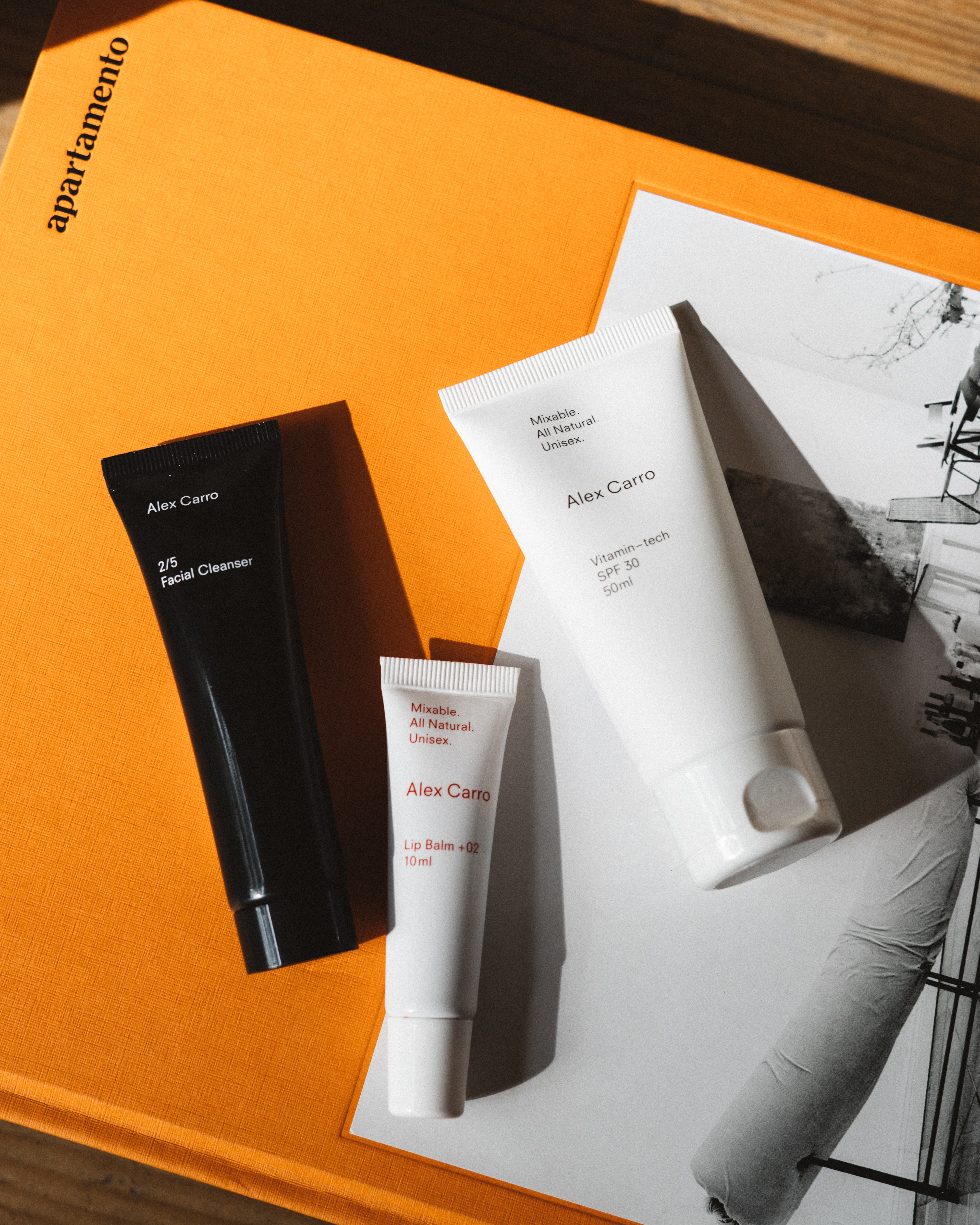 Three white and black cosmetic tubes from Alex Carro are displayed on an orange book cover and a blurred black and white photo.