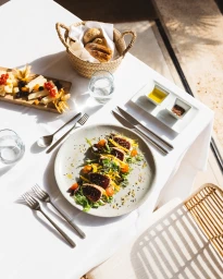 High angle view of a dining table with a seared tuna salad, bread basket, cheese board, and cutlery in bright sunlight.