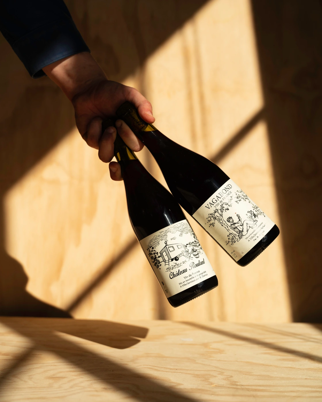 A hand holding two bottles of natural wine diagonally against a light plywood surface bathed in diagonal sunlight and shadows.