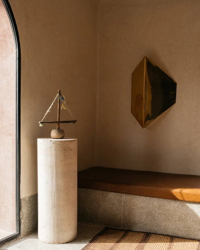 Stone pedestal with a miniature wooden boat sculpture beside a geometric brass wall piece in a warm sunlit interior.