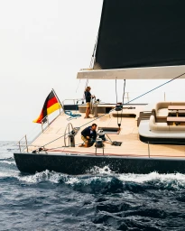 A woman steers a modern luxury yacht with a large black sail and German flag across deep blue ocean water alongside a crew member on deck.