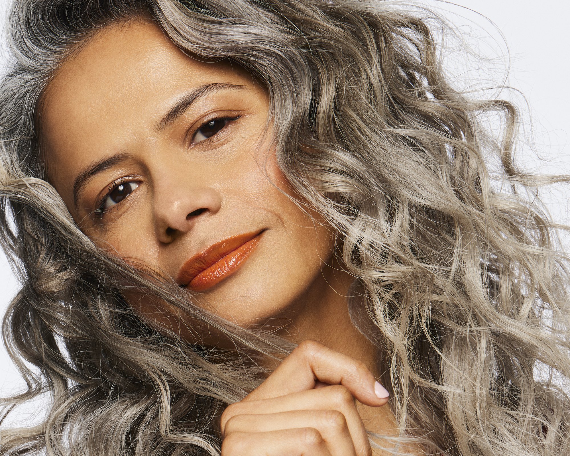 Close-up portrait of a mature woman with silver curly hair, warm skin tone, and glossy orange lipstick against a white background.