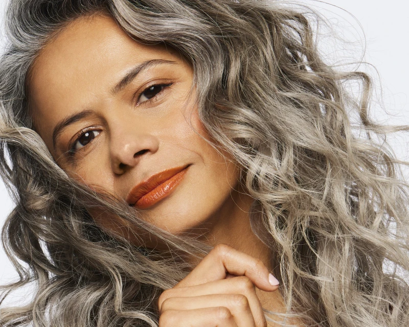 Close-up portrait of a mature woman with silver curly hair, warm skin tone, and glossy orange lipstick against a white background.