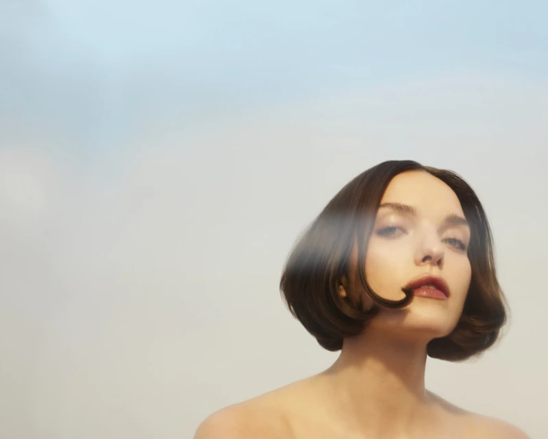 Portrait of a woman with short brunette hair and bare shoulders against a soft background. Light from an unseen source highlights her facial features.