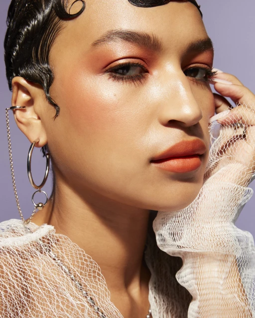 Close-up portrait of a woman with orange makeup and a net shawl. Features hoop earrings and stylized dark curly hair.
