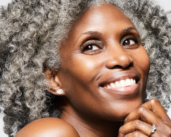 Close-up portrait of an older Black woman with curly grey hair smiling against a white seamless background.