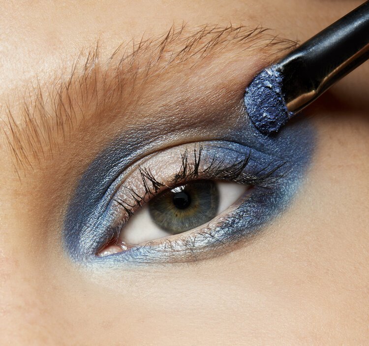 Extreme close-up of blue metallic eyeshadow being applied with a makeup brush to an eye with dark lashes and brow
