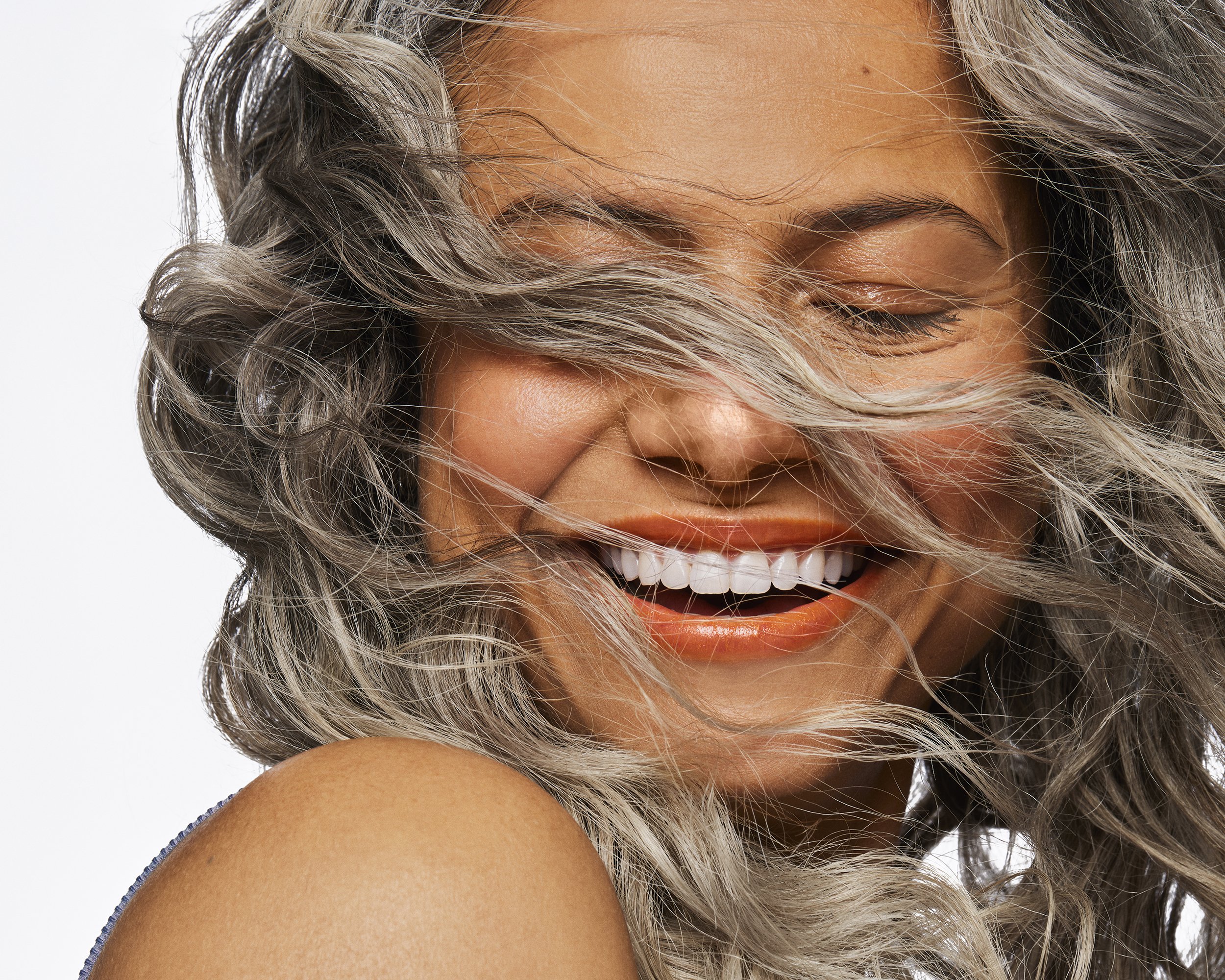 Close-up portrait of a laughing adult woman with silver, wavy hair against a clean white background; showing shoulder, and closed eyes.