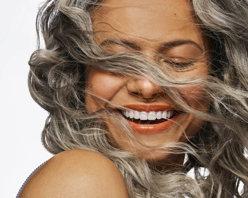 Close-up portrait of a laughing adult woman with silver, wavy hair against a clean white background; showing shoulder, and closed eyes.