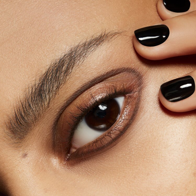 Close-up studio shot of an adult female's eye with brown iris, arched eyebrow, and manicured fingernails painted with black nail polish.