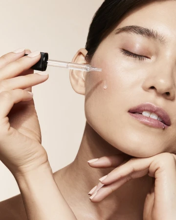 Close-up of adult female applying liquid from clear pipette to her cheek, with a beige background, promoting skincare and beauty.