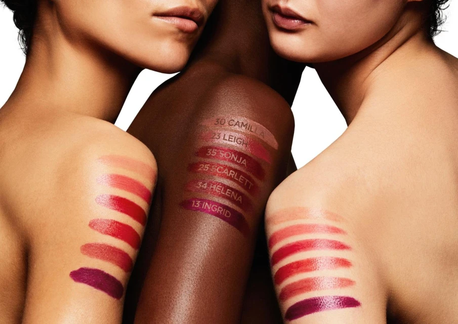 Close-up of three women with varying skin tones showcasing lipstick swatches on their shoulders in a beauty and cosmetics editorial.