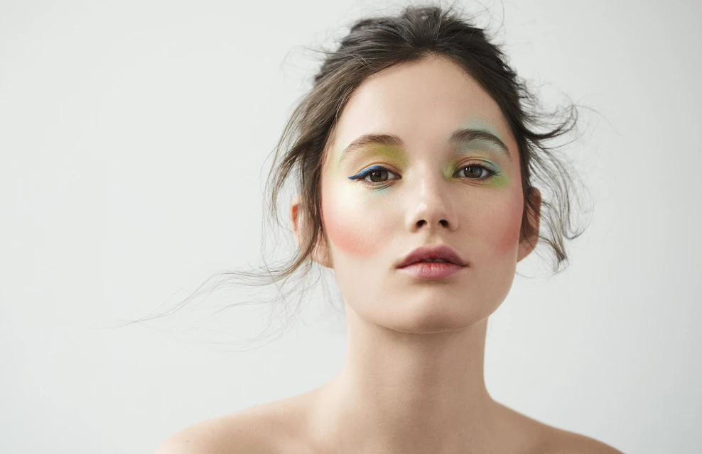 Close-up of a woman with colorful makeup on her eyes and cheeks, shot in a photo studio with a clean, white background.