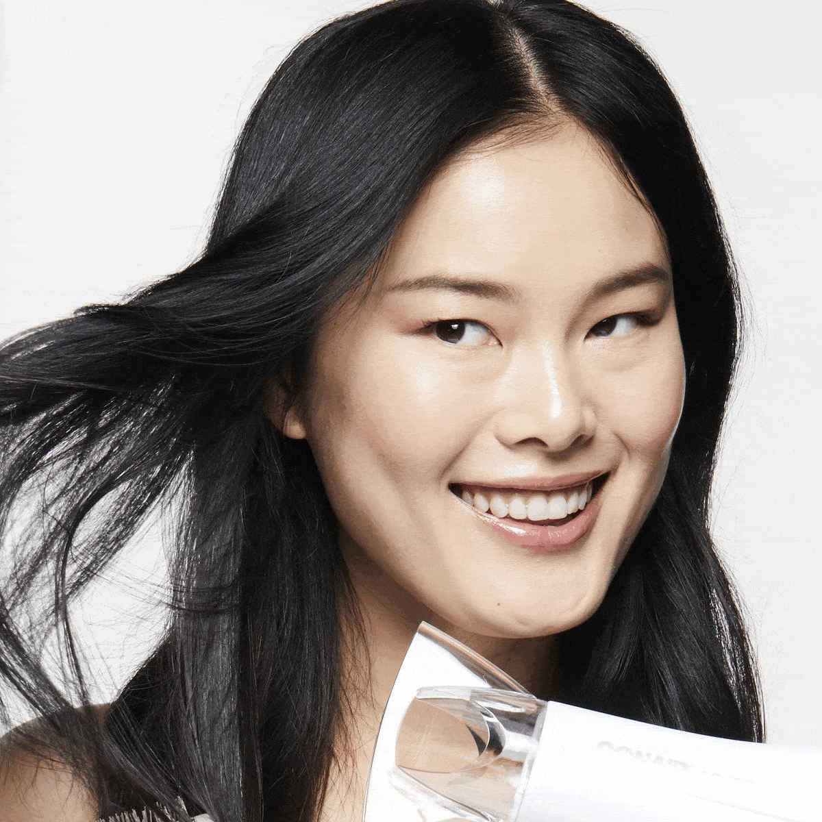 Close-up of a smiling Asian woman with long black hair, using a white hair dryer against a white background in a beauty shot.