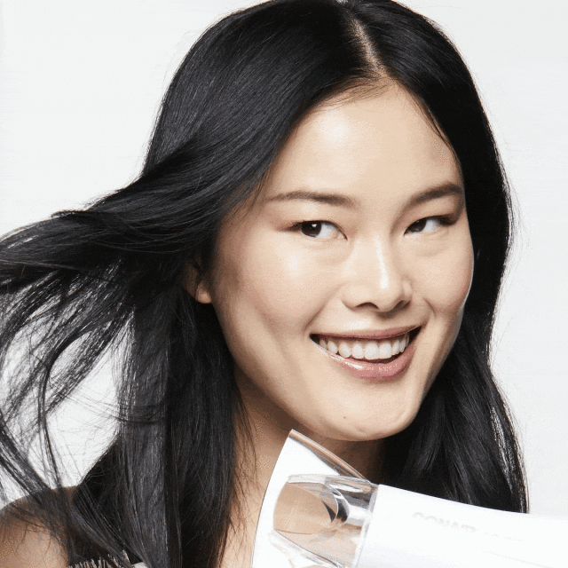 Close-up of a smiling Asian woman with long black hair, using a white hair dryer against a white background in a beauty shot.