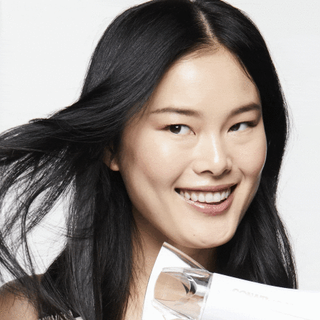 Close-up of a smiling Asian woman with long black hair, using a white hair dryer against a white background in a beauty shot.