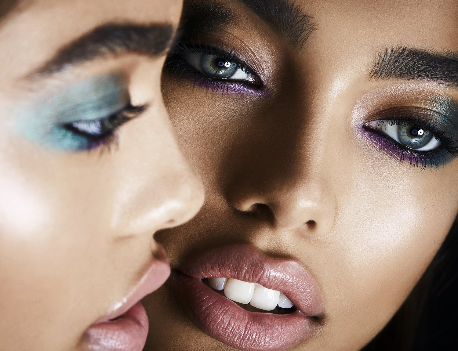 Close-up of two diverse women with colorful eye makeup. One woman has teal makeup, the other with darker teal and purple tones.