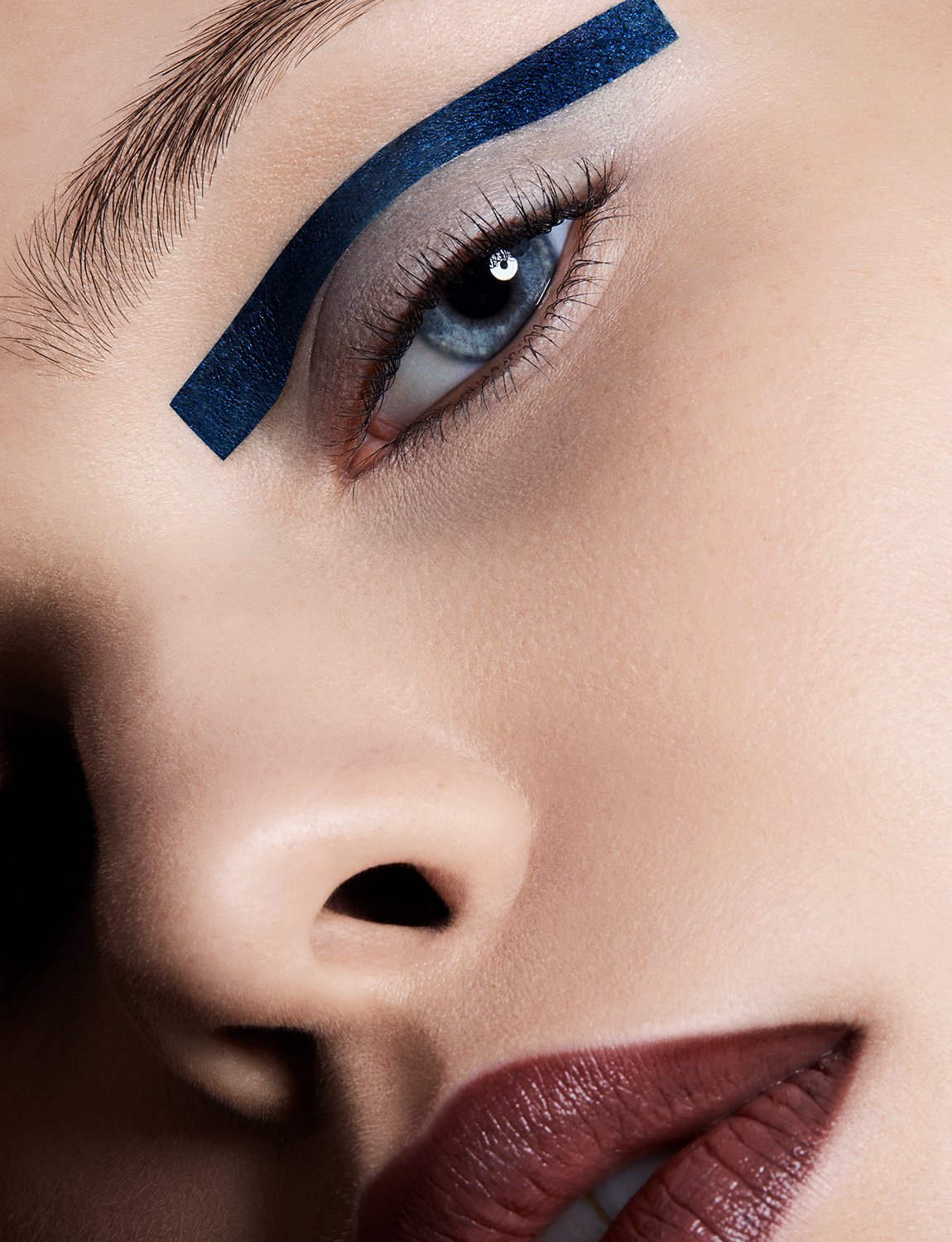 Close-up beauty shot of a woman's face with blue eye makeup and dark lipstick, featuring fair skin and an intense gaze.