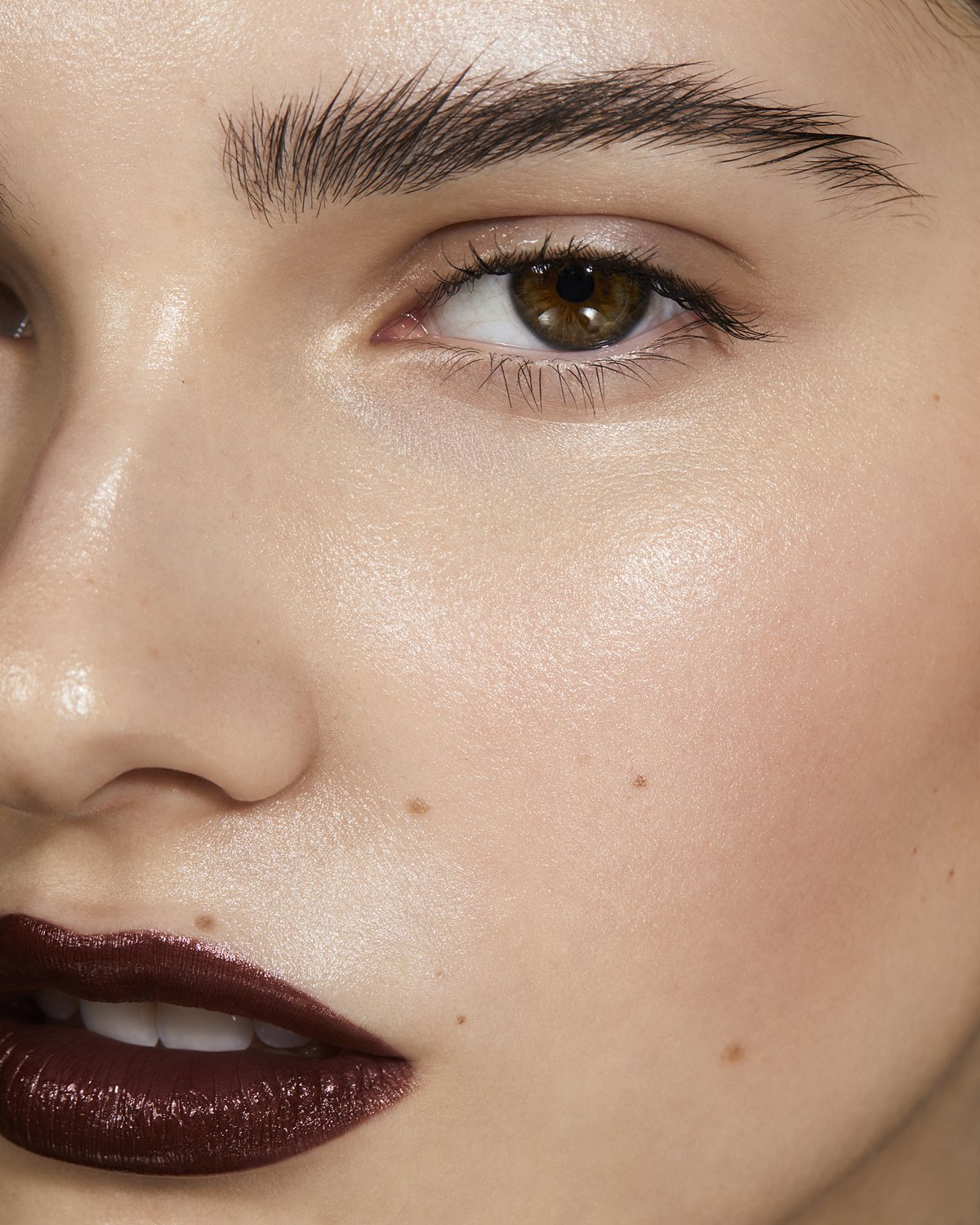 Close-up of a woman's face with glossy, dark red lipstick, prominent eyebrows, and smooth, glowing skin, emphasizing makeup and beauty.