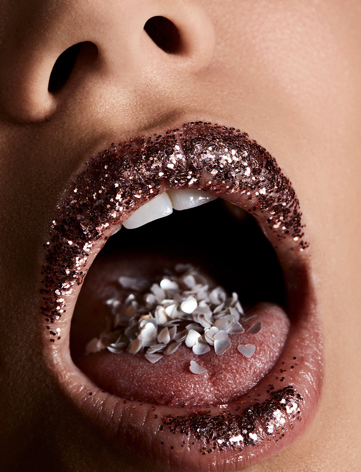 Woman's open mouth with rose gold glitter lipstick and tongue covered in pearlescent heart-shaped objects in a close-up shot.