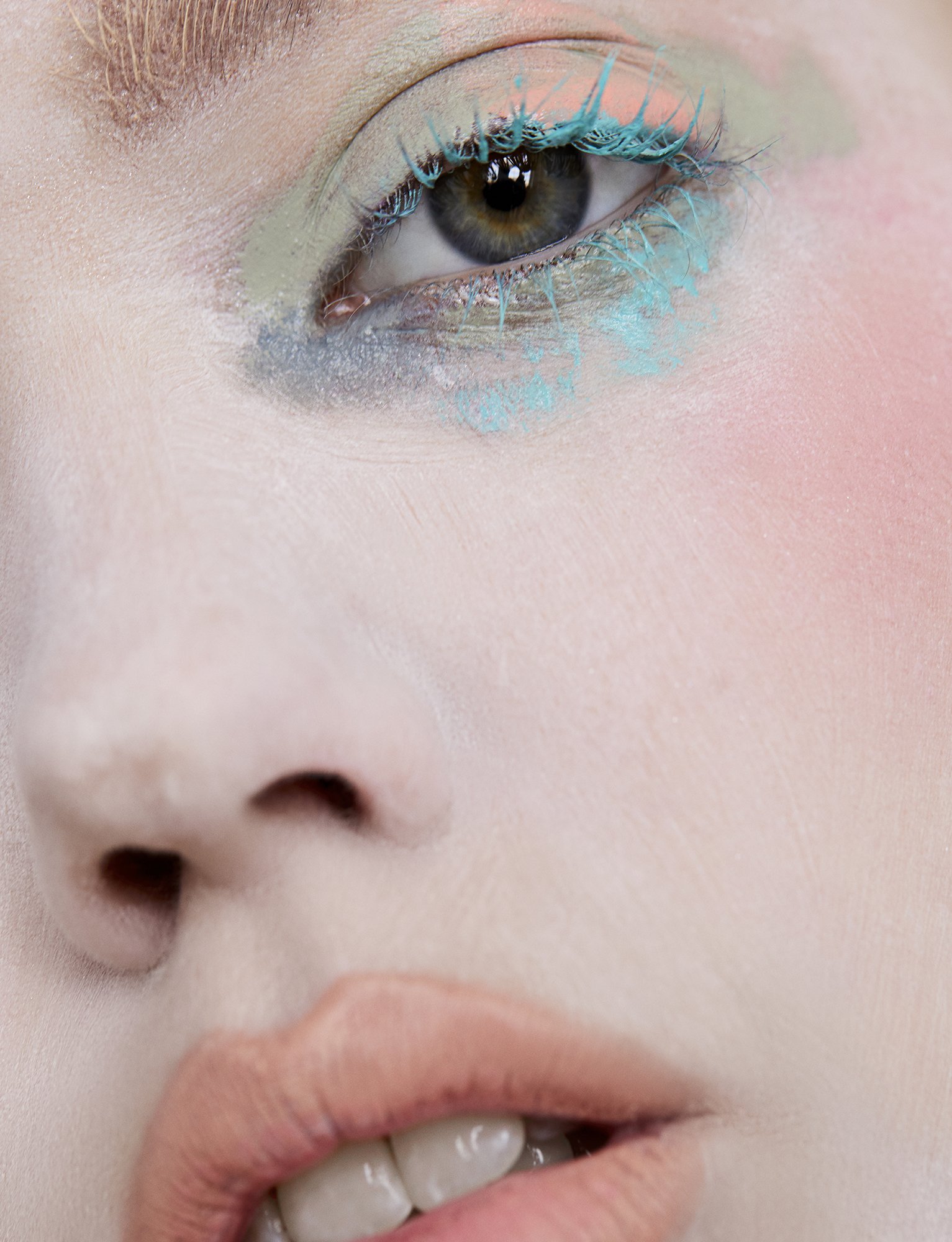 Close-up of a woman's face with teal and green eye makeup, pale skin, and glossy lips. Beauty shot emphasizing makeup and skin tones.
