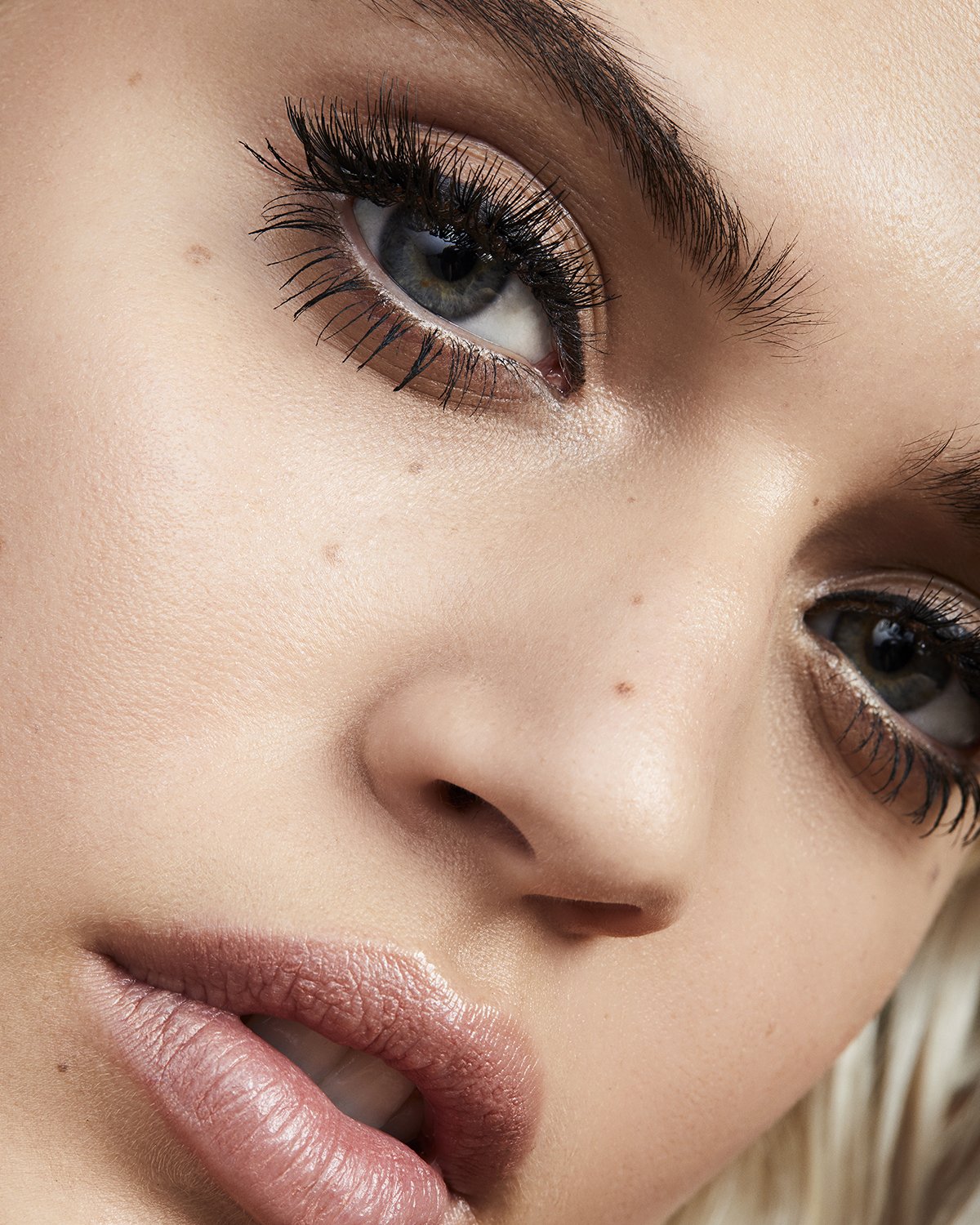 Close-up beauty shot of a young adult female face featuring blue eyes with mascara, light skin, and nude lipstick.