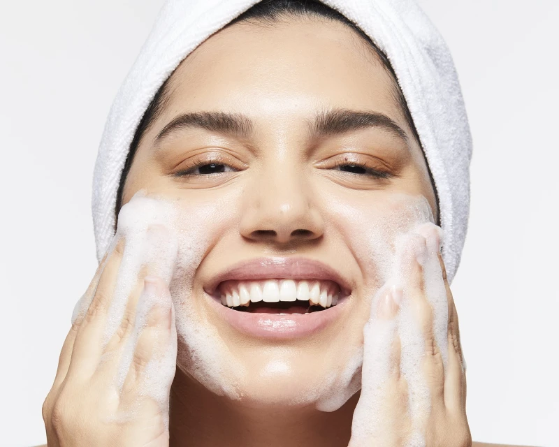 Smiling woman washes her face with foaming cleanser while wearing a white towel wrapped around her head against a white backdrop.