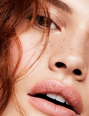Close-up portrait of a woman with red hair, pale skin, freckles, and soft pink lips under direct lighting.