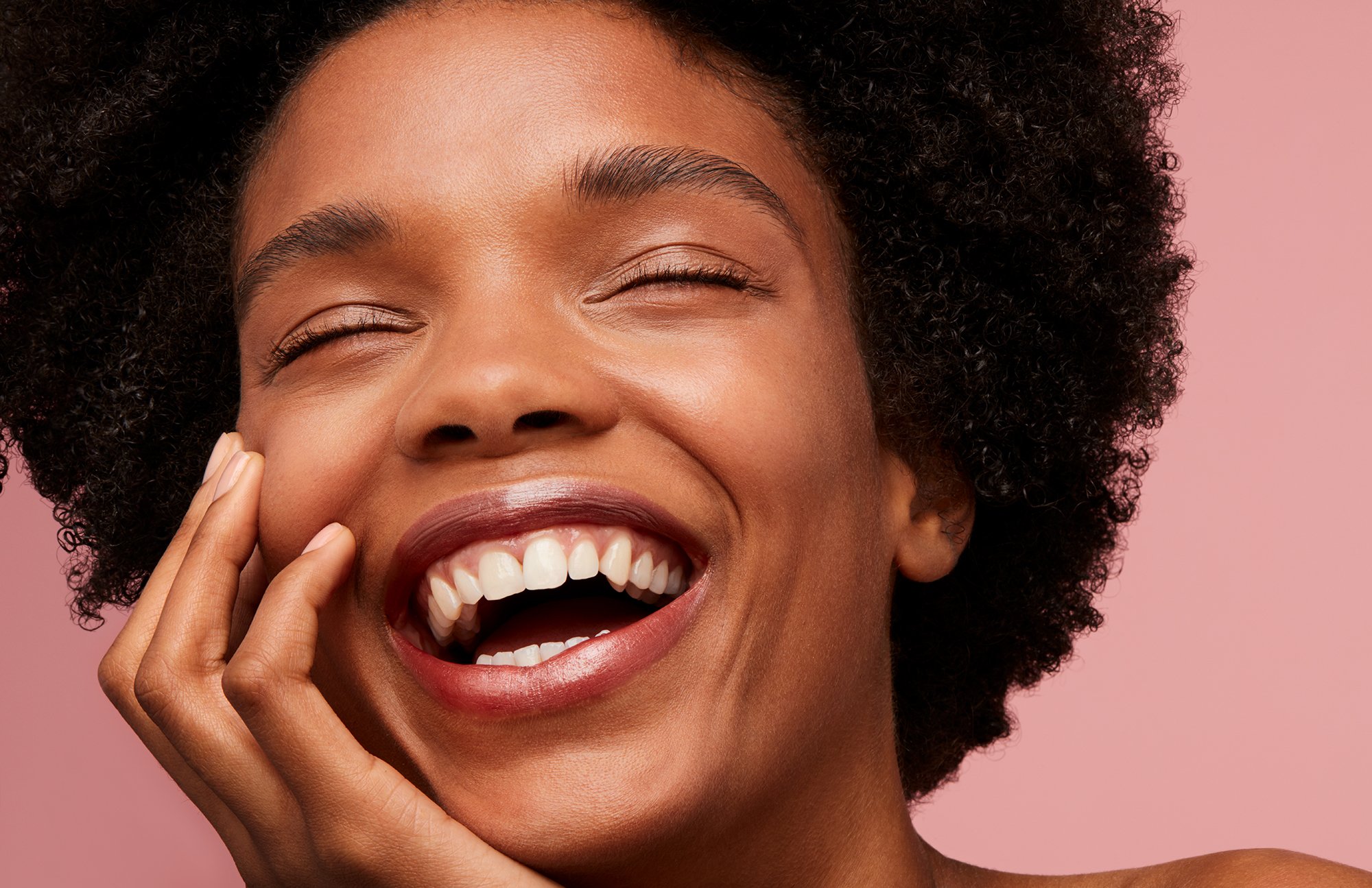 Close-up portrait of a Black woman laughing with eyes closed and hand near chin against a pink background