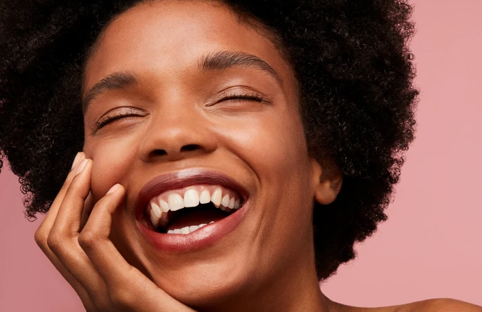 Close-up portrait of a Black woman laughing with eyes closed and hand near chin against a pink background