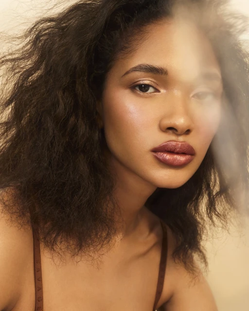 Close-up portrait of an adult woman with brown curly hair, soft, natural makeup, and a brown top against a cream background in a photo studio.
