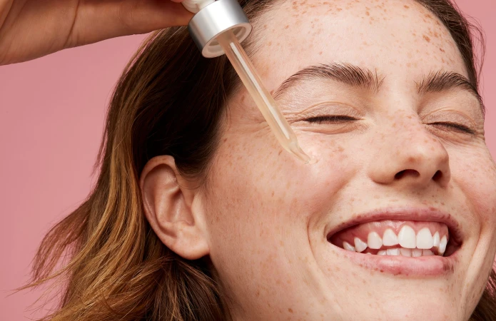 Close-up of a smiling woman with heavy freckles having serum dropped onto her cheek from a glass pipette.