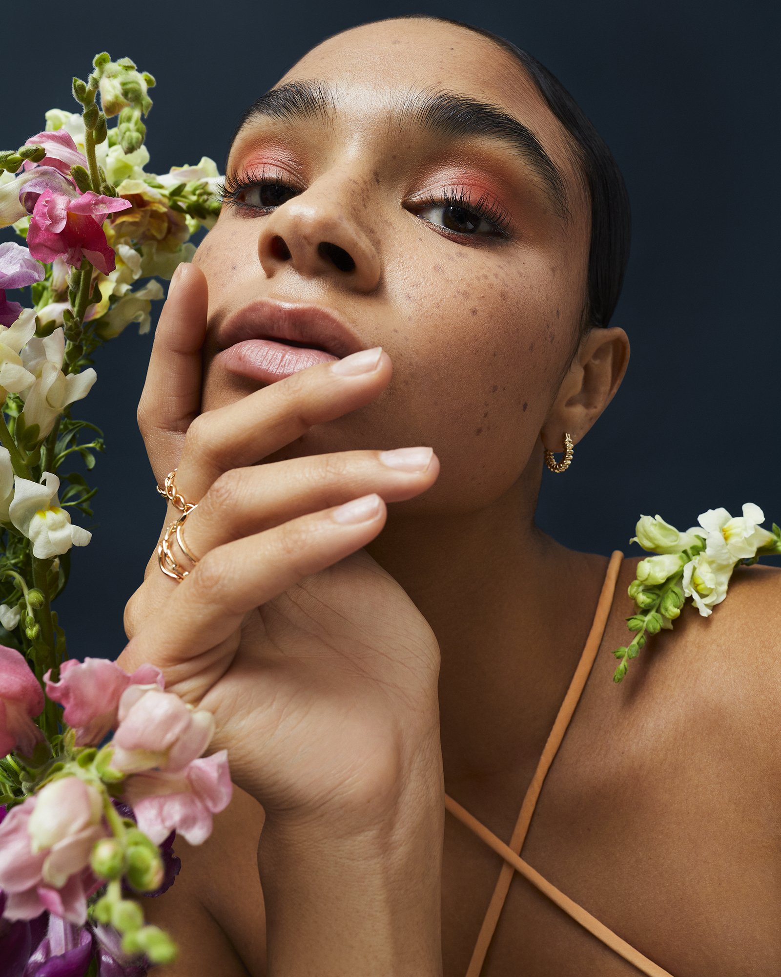 Close-up portrait of a woman with freckles and coral eye makeup, adorned with flowers, set against a dark blue background.