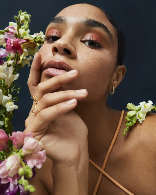 Close-up portrait of a woman with freckles and coral eye makeup, adorned with flowers, set against a dark blue background.
