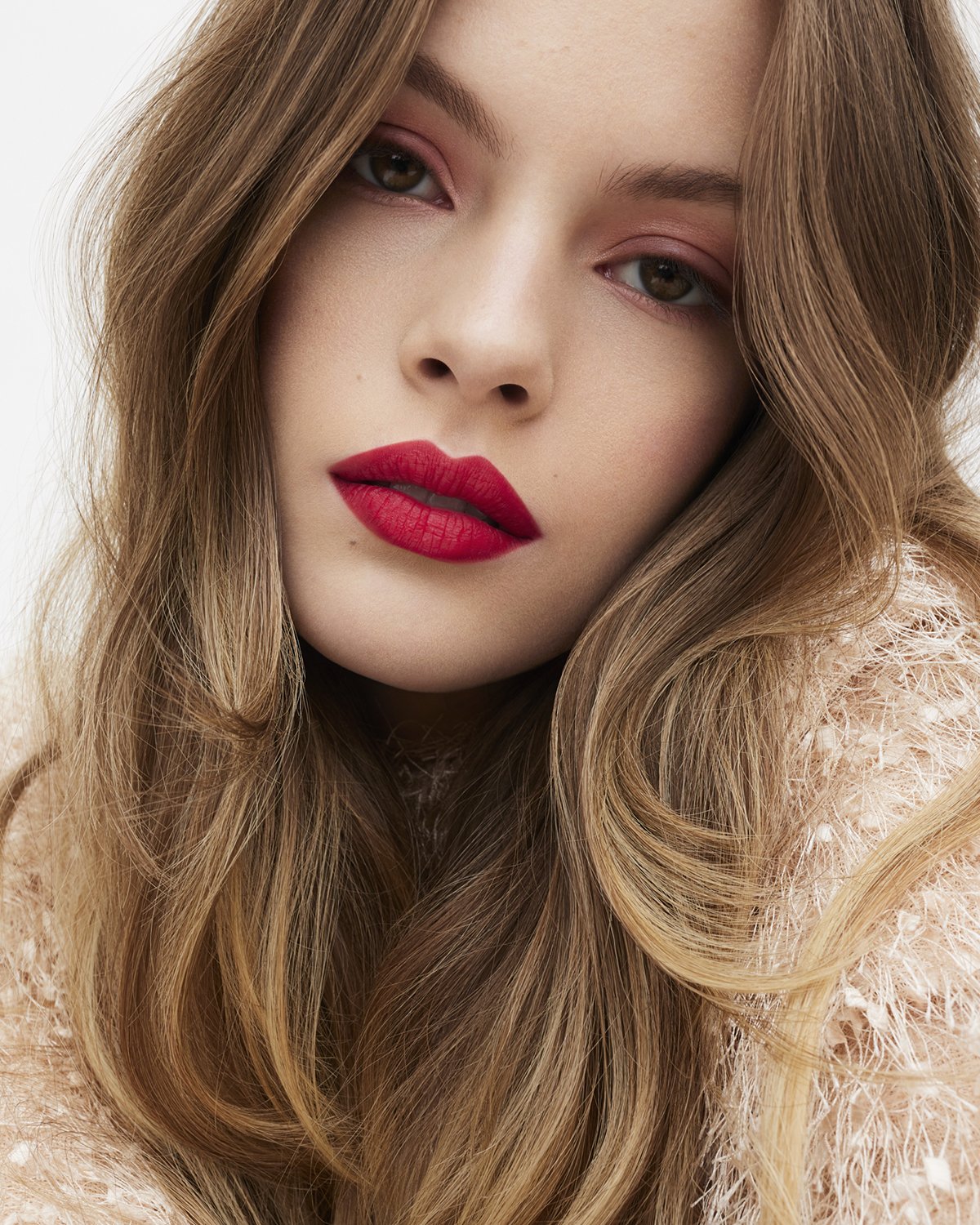 Close-up studio portrait of a young woman with long wavy auburn hair, red lipstick, and red eyeshadow against a clean white background.