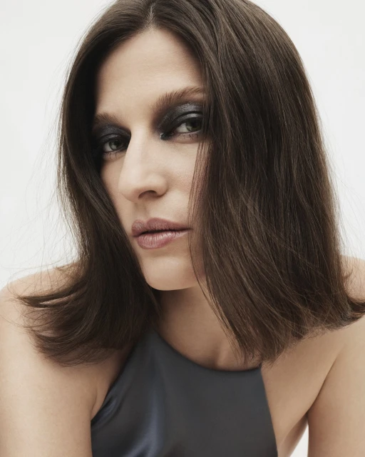 Close-up portrait of an adult woman with dark hair and smokey eye makeup, wearing a grey-blue halter top against a white background.