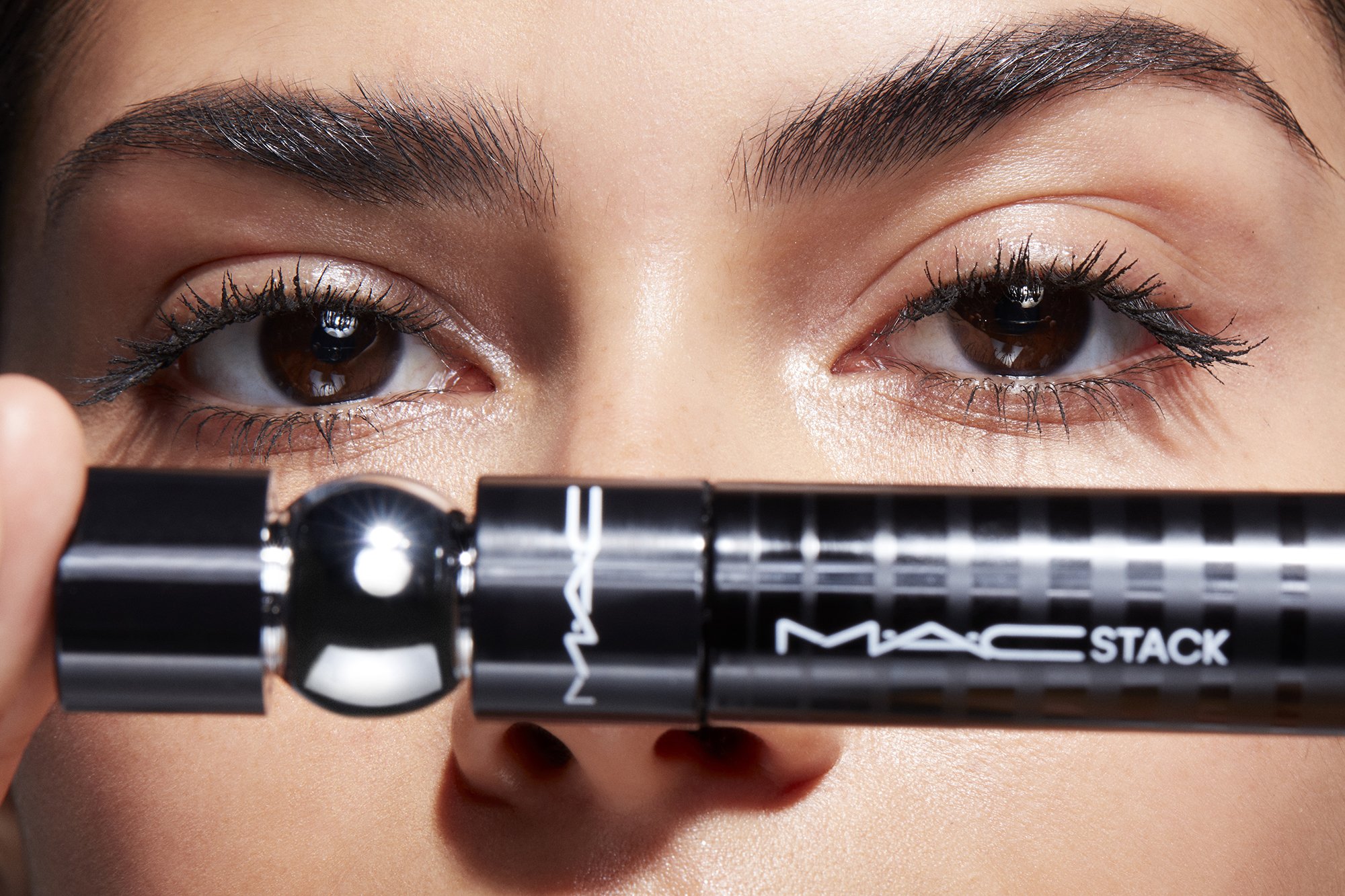 Close-up of woman's face with brown eyes and mascara, holding black MACStack mascara product in front of her nose in studio lighting.